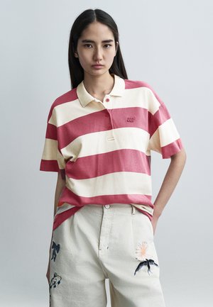 Tricou polo cu dungi roșii și crem, mâneci scurte, cu guler și trei nasturi. Are un logo mic brodat pe piept.