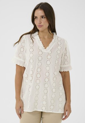 Femme portant un chemisier blanc en dentelle œillet avec des manches courtes à volants et un col en V, associé à un pantalon beige, regardant sur le côté.