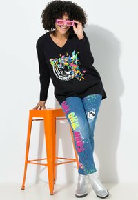 Camiseta negra de manga larga con gráfico de tigre, jeans azules con manchas coloridas y texto, y botines metálicos plateados. Taburete naranja.