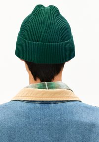 Beanie verde a coste con orlo arrotolato e pattern texturizzato, indossato al contrario da una persona con capelli scuri, sovrapposto a una camicia a quadri.