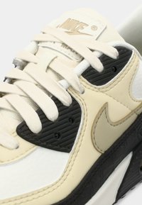 Nike sneaker, mis kogub valget, kreemi ja musta materjali segu. Tekstuuriga paneelid ja siledad paelad, millel on logo keelega ja külgedel.
