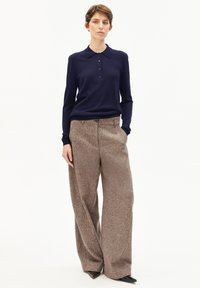 Dunkelblaues Polohemd mit langen Ärmeln, Kragen und Knopfleiste, kombiniert mit weiten, braunen, strukturierten Hosen und spitzen schwarzen Schuhen.