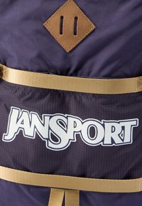 Μωβ υφασμάτινο σακίδιο πλάτης με ανοιχτό καφέ διακοσμητικό ρόμβο και χρυσά λουριά, με μεγάλο λευκό λογότυπο "JANSPORT" στο μπροστινό μέρος.