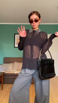 Persona con gafas de sol tintadas, camisa negra transparente con cuello estampado, pantalones grises de pierna ancha, sosteniendo un bolso negro en interiores.