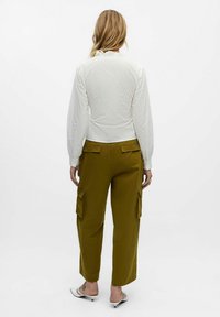 Femme aux cheveux blonds se tient de dos, portant un blouse blanche à manches longues, un pantalon cargo vert olive et des chaussures blanches à petits talons.