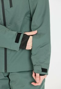 Grüne, strukturierte Jacke mit einem Reißverschluss, verstellbaren Manschetten und einer versteckten Tasche. Mit schwarzem Beschlag und einer taillierten Passform.