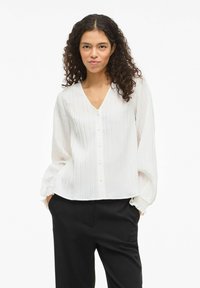 Hvid bluse med lodrette plisseringer, V-udskæring og knaplukning foran. Har lange, vide ærmer med elastiske manchetter. Bæres med sorte bukser.