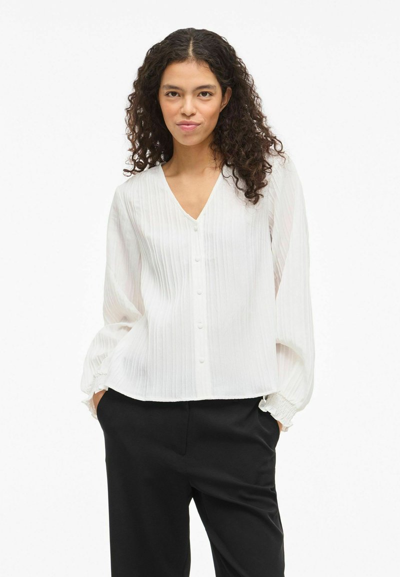 Hvid bluse med lodrette plisseringer, V-udskæring og knaplukning foran. Har lange, vide ærmer med elastiske manchetter. Bæres med sorte bukser.