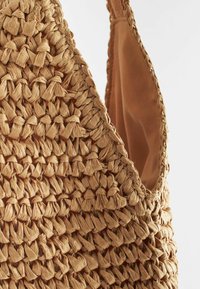 Wykonana z plecionego beżowego słomy torba z teksturowaną powierzchnią i gładkim brązowym wykończeniem. Posiada mocny pasek oraz luźny fason.