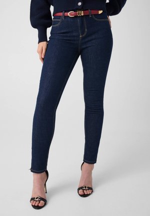 Femme portant un jean skinny bleu foncé, une ceinture rouge, des sandales noires à talons avec un détail doré, et un pull bleu marine.