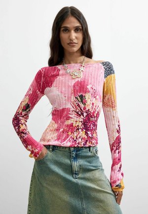 Donna che indossa un top colorato a maniche lunghe con motivo floreale e textura a costine, gonna di jeans blu-verde e una collana d'argento floreale.