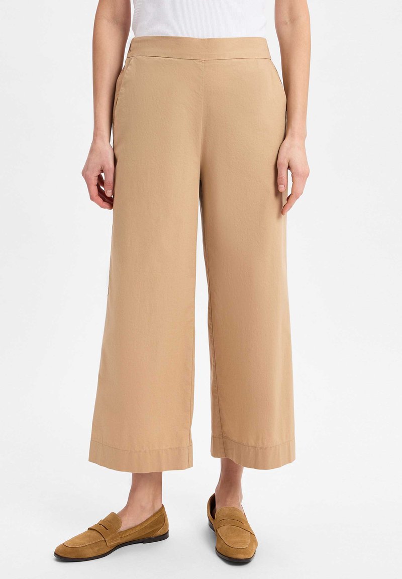 Beige weite Hose mit glatter Textur, Fronttaschen und sauberem Saum, kombiniert mit hellbraunen Slippern.