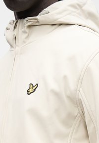 Kreminā krāsas jaka ar gludu tekstūru, kapuci un nelielu melni-zeltainu izšūtu putnu logo uz kreisās krūšu puses.