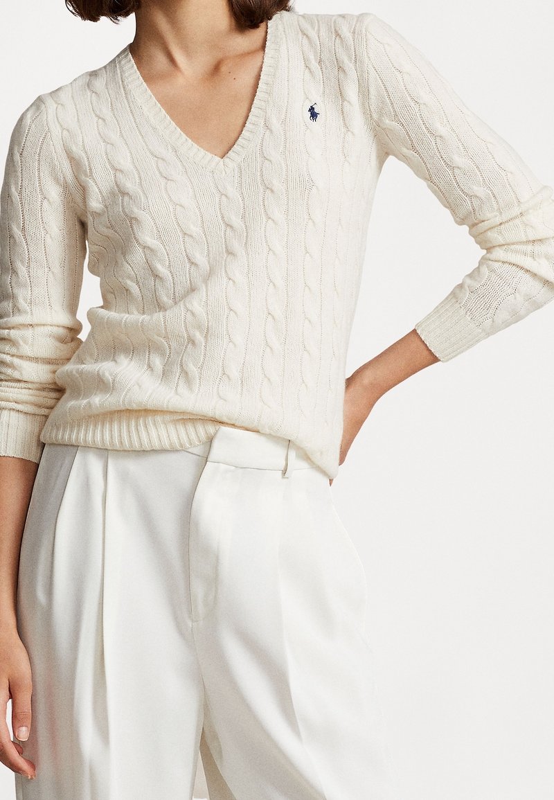 Femme portant un pull torsadé col V écru avec logo bleu et pantalon blanc plissé taille haute, main posée sur la hanche.
