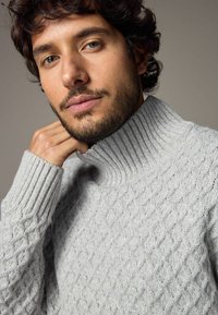 Hellgrauer Strickpullover mit hohem geripptem Kragen und Rautenmuster-Textur. Nahaufnahme des Stoffes und der Detailansicht des Ärmels.