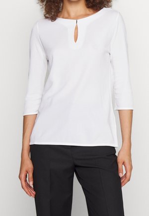 Femme portant un haut blanc à manches longues avec un petit détail en forme de trou à l'avant et un pantalon noir ajusté, sur un fond uni.