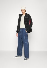 Svart dunjakke med teksturert stoff, beige t-skjorte, løse blå cargo-jeans og grå joggesko med hvite striper. Lut hue på hodet.