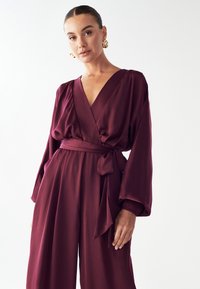 Burgundska oblačila z dolgimi rokavi, V-izrezom in zavezanim pasom. Mehka, svilnata tekstura z elastičnimi manšetami za dodaten poudarek.