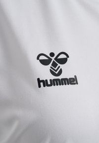 Biała sportowa koszulka z czarnym logo stylizowanej pszczoły nad nazwą marki "hummel" w pogrubionym, czarnym tekście. Gładka, syntetyczna tkanina.