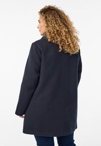 Navy wool coat with a straight cut, back vent detail, and long sleeves; textured material; mid-thigh length.