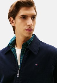 Marine corduroy jas met rits en spitse kraag, gelaagd over een groen geruit overhemd met witte en blauwe accenten. Heeft logodetail.