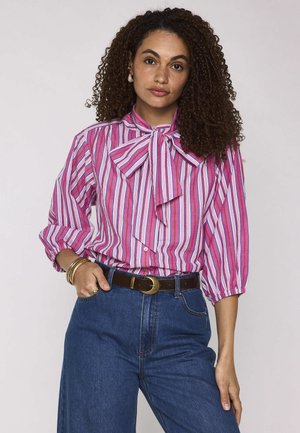 Vrouw met krullend haar, draagt een roze-wit gestreept blouse met strikjeskraag, blauwe spijkerbroek en gouden armbanden, staand met één hand in de zak.