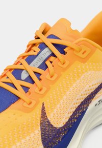 Πορτοκαλί και μωβ αθλητικό παπούτσι με υφασμάτινο άνω μέρος με υφή, σχέδιο κορδονιών και ορατό λογότυπο Nike swoosh στο πλάι.