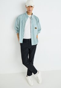 Chemise à rayures bleu clair à manches longues, t-shirt blanc, pantalon noir, baskets blanches et casquette crème. Présente un patch logo sur la chemise.