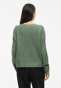 Pull en maille épaisse de couleur vert atténué, avec un col rond, des épaules tombantes, des détails de poignets côtelés, et une coupe décontractée qui tombe à la taille.