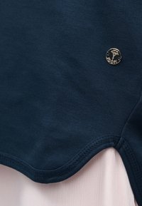 Υφασμα σε ναυτικό μπλε με καμπυλωτή ραφή πάνω σε ανοιχτό ροζ ύφασμα, που διαθέτει μικρό στρογγυλό μεταλλικό σήμα με σχεδιασμό λουλουδιού και κείμενο.