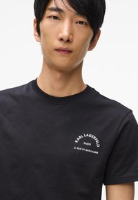 Melnas kokvilnas t-krekls ar apaļu izgriezumu. Iekļauj baltu apdrukātu tekstu: "KARL LAGERFELD PARIS 21 RUE ST-GUILLAUME" kreisajā krūtīs.