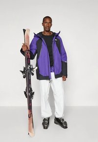 Lila-schwarze Skijacke mit Kapuze, schwarzer Baselayer, weiße Skihose, hält Skier mit Bindungen. Glatte Textur, modernes Design.