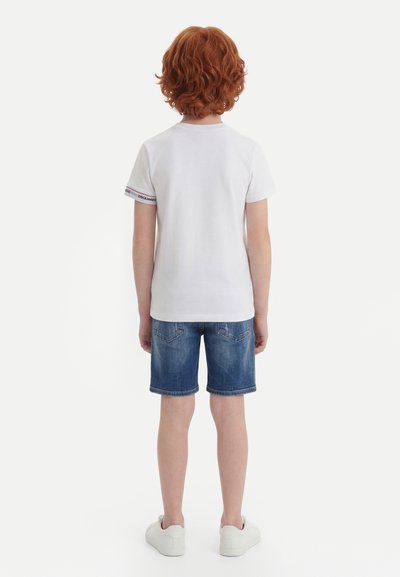 Bambino con capelli rossi che indossa una maglietta bianca, pantaloncini di jeans e scarpe da ginnastica bianche, in piedi di spalle su uno sfondo neutro.