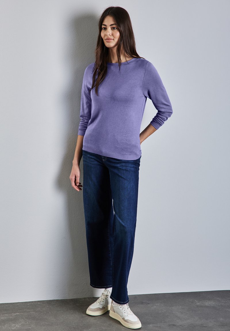 Ljuslila tröja med trekvartsärmar, tillsammans med mörkblå vida jeans och vita sneakers. Slät textur och avslappnad design.