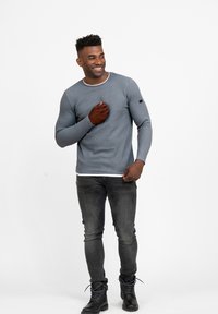 Graues, geripptes Langarmshirt mit weißem Kragen, kombiniert mit dunkelgrauen Jeans und schwarzen Stiefeln. Auf dem Arm befindet sich ein kleines Logo.