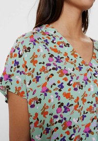 Blusa a stampa floreale verde menta, con maniche corte, scollatura a V e un tessuto leggero decorato con motivi arancioni, viola e marroni.