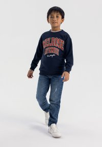 Felpa della Marina con grafica "MELROSE AVENUE" in rosso e bianco, abbinata a jeans chiari strappati e sneakers bianche.