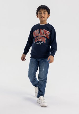 Marineblauwe sweatshirt met de opdruk "MELROSE AVENUE" in rood en wit, gecombineerd met lichtblauwe distressed jeans en witte sneakers.