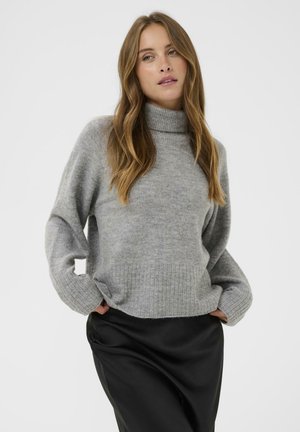 Woman with long wavy hair wearing a gray turtleneck sweater and black skirt, standing with hands in pockets against a plain background.