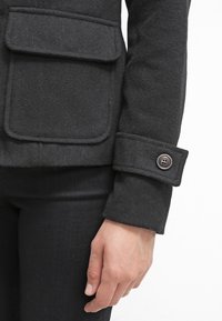 Veste en laine noire avec une grande poche à rabat, un poignet à bouton et une finition texturée. La manche s'étend jusqu'au poignet avec un pli.