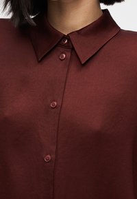 Rødbrun skjorte med knapper, der har en spids krave, fem matchende knapper og et glat, let tekstureret stof.