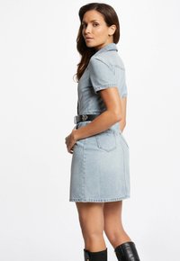 Denim-paita-asu vaaleansinisessä, kiinteässä muotoilussa, lyhyet hihat, vyötärö vyöllä ja taskut takana. Musta vyökorostus.