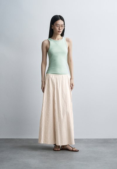 Femme aux longs cheveux noirs et lunettes porte un haut sans manches vert menthe, un pantalon beige à larges jambes rayé et des sandales marron, debout à l'intérieur.