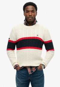 Maglione a trecce color crema, con strisce orizzontali rosse e blu navy. Indossato sopra una camicia a quadri, con collo e polsini a coste.
