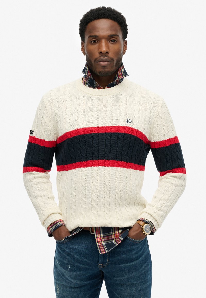 Maglione a trecce color crema, con strisce orizzontali rosse e blu navy. Indossato sopra una camicia a quadri, con collo e polsini a coste.