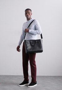 Svart läder tote-väska med texturerat mönster, havremanschetter och justerbar axelrem. Modellen bär en ljusblå skjorta med långa ärmar och vinröda byxor.
