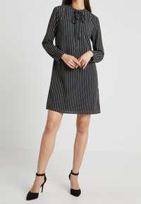 Vestito a righe nere con maniche lunghe, chiusura con bottoni frontale e dettaglio del collo a cravatta. Tessuto morbido, vestibilità ampia,lunghezza sopra al ginocchio, abbinato a tacchi neri.