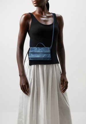 Pequeño bolso cruzado de cuero azul con un diseño estructurado, que presenta dos solapas frontales y un asa en la parte superior. Combinado con una camiseta sin mangas negra y una falda blanca.