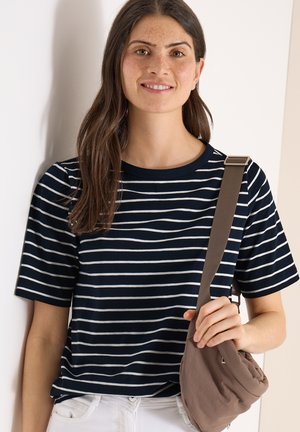 Junge Frau mit langen braunen Haaren, die ein marineblau-weiß gestreiftes Shirt trägt, den Riemen einer braunen Umhängetasche hält und sanft lächelt.