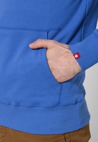 Sudadera de algodón azul con un bolsillo canguro y puños acanalados. Presenta una pequeña etiqueta roja en el puño. Textura suave con un corte relajado.